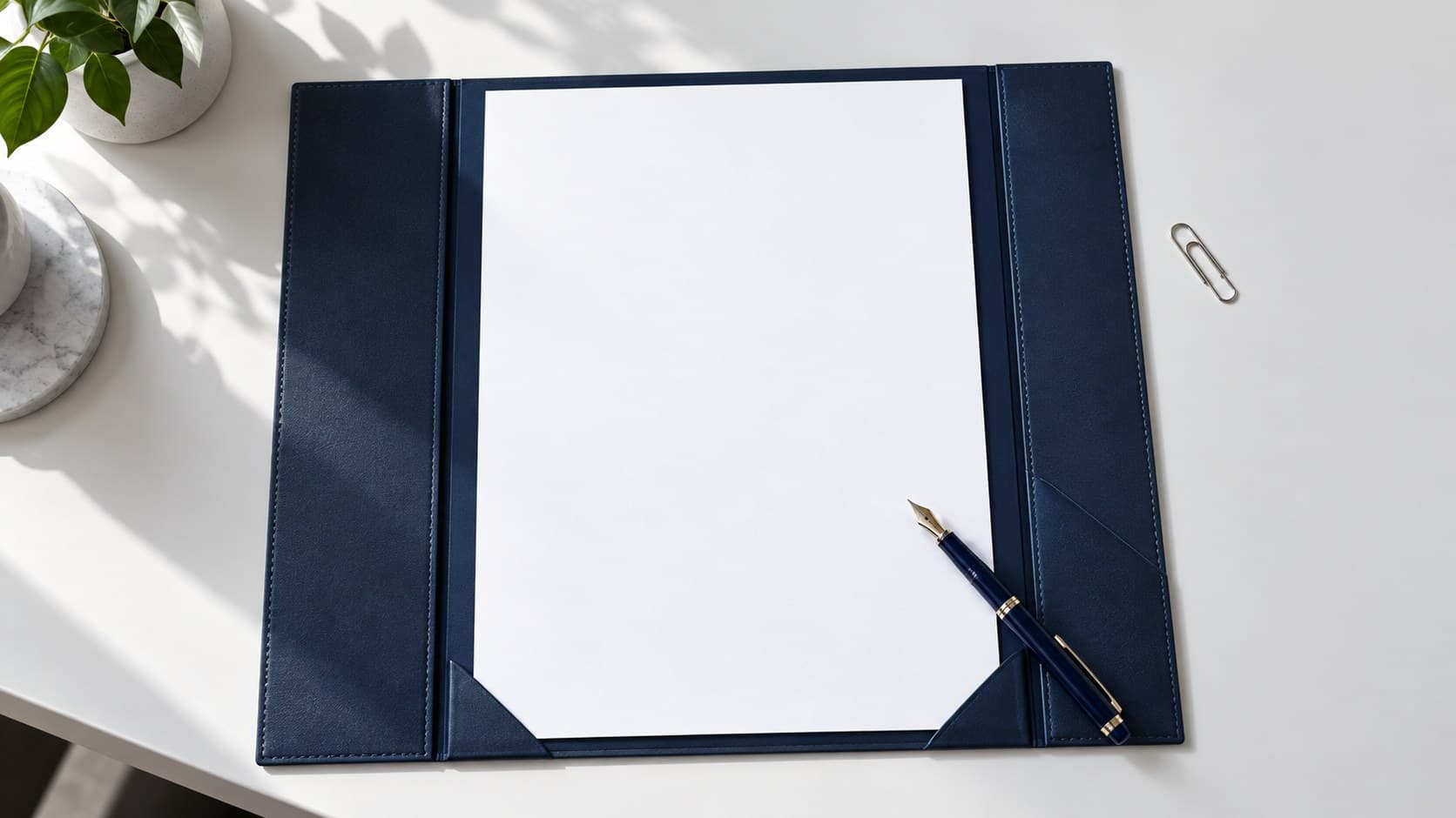 A blank sheet sits inside an open navy document folder on a white desk, beside a blue fountain pen and a paper clip.